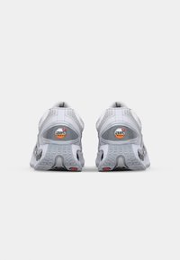 Bijele atletske cipele s mrežastim gornjim dijelom i gumeni potplat. Sadrže Air Max logo, teksturalne uzorke sa strane i kontrastne crvene akcentne detalje na potplatu.