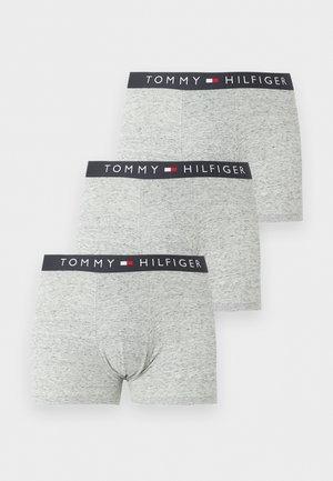 Shirtloos mannelijk model staat blootsvoets en draagt grijze Tommy Hilfiger boxershorts met zwarte tailleband op een witte achtergrond.