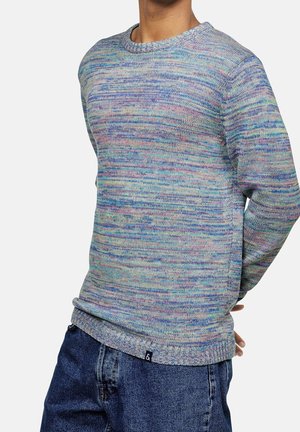 Persona vistiendo un suéter de punto multicolor en tonos pastel y jeans azules, de pie con las manos detrás de la espalda, sobre un fondo liso.