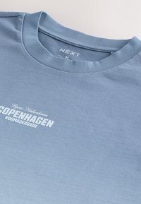 Jasnoniebieska koszulka z okrągłym dekoltem, z białym napisem "Byen København COPENHAGEN KØBMAGERGADE" oraz metką z rozmiarem "NEXT M" wewnątrz kołnierza.
