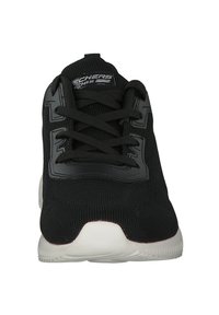 Black athletic shoe with mesh upper, rounded toe, and flat laces. Features a textured sole and branding on the tongue and side.