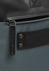 Schwarze Leder- und graue Stoff-Rucksacksektion mit einem Reißverschluss, strukturiertem Material und einem schwarzen Lederriemen mit silberfarbenen Hardware-Details.