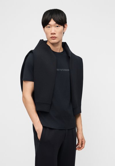 Νεαρός άνδρας που φοράει μαύρο μπλουζάκι και παντελόνι με μαύρο φούτερ ριγμένο στους ώμους, στέκεται μπροστά σε απλό ανοιχτό φόντο.