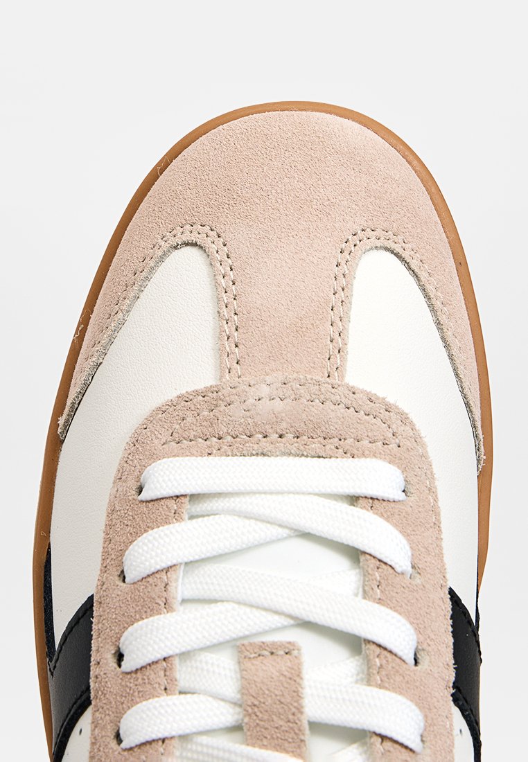 Beige Wildleder- und weiße Leder-Sneaker mit brauner Gummisohle, weißen Schnürsenkeln und schwarzem Seitenstreifen, von oben nah aufgenommen.