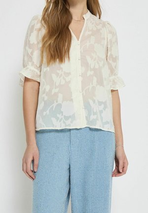 Nainen pukeutuneena läpikuultavaan valkoiseen kukkakuviolliseen paitaan, jossa on lyhyet puhvimaiset hihat, ja vaaleansinisiin, kuvioituihin, korkeavyötäröisiin housuihin.