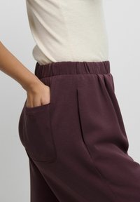 Pantaloni bordo din amestec de bumbac, cu talie elastică, buzunar lateral și croială lejeră, expuși alături de un top bej mulat.