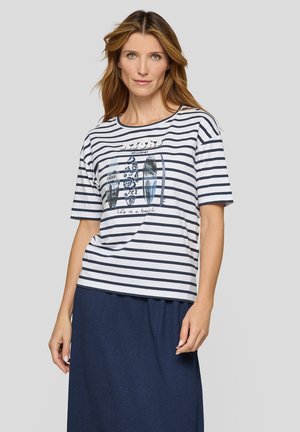 Frau mit langen hellbraunen Haaren, die ein weißes und marineblau gestreiftes T-Shirt mit Surfbrett-Grafiken und einen marineblauen Rock vor einem grauen Hintergrund trägt.