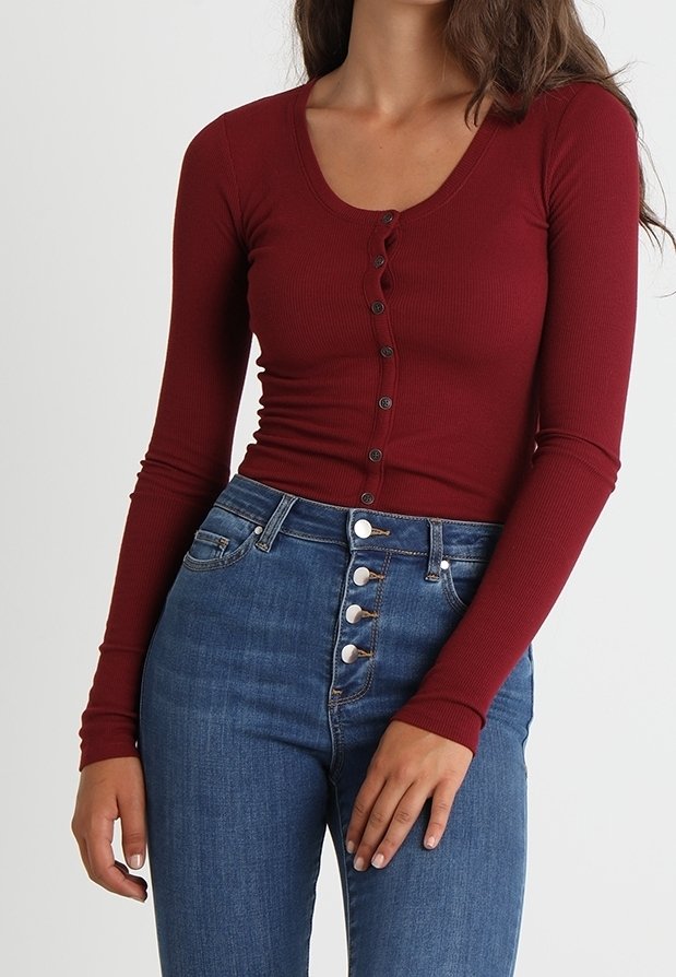 Pitkähihaista viininpunaista ribbipaitaa, jossa on syvä kaula-aukko ja nappikiinnitys edessä, yhdistettynä korkeavyötäröisiin sinisiin farkkuihin.