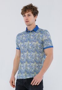 Camisa polo de manga corta con un patrón abstracto en azul y verde, cuello y puños azules, tapeta de botones y tela de algodón texturizada.