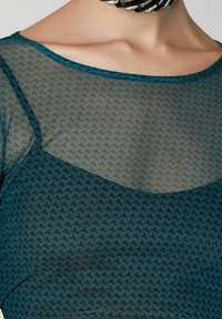 Tyrkysový síťovaný top s houndstooth vzorem, s kulatým výstřihem a tenkými ramínky. Látka je průhledná, což dodává texturu designu.