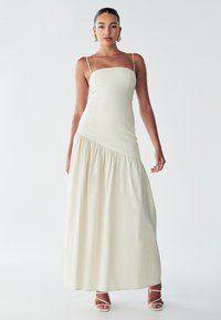 Robe maxi en tissu crème avec un corsage ajusté et une jupe fluide à volants. Comprend des fines bretelles spaghetti et une texture lisse.