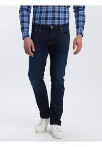 Muž oblečený v tmavě modrých slim-fit džínách, bílých teniskách a modré kostkované košili s dlouhým rukávem, stojící na jednolitém pozadí.