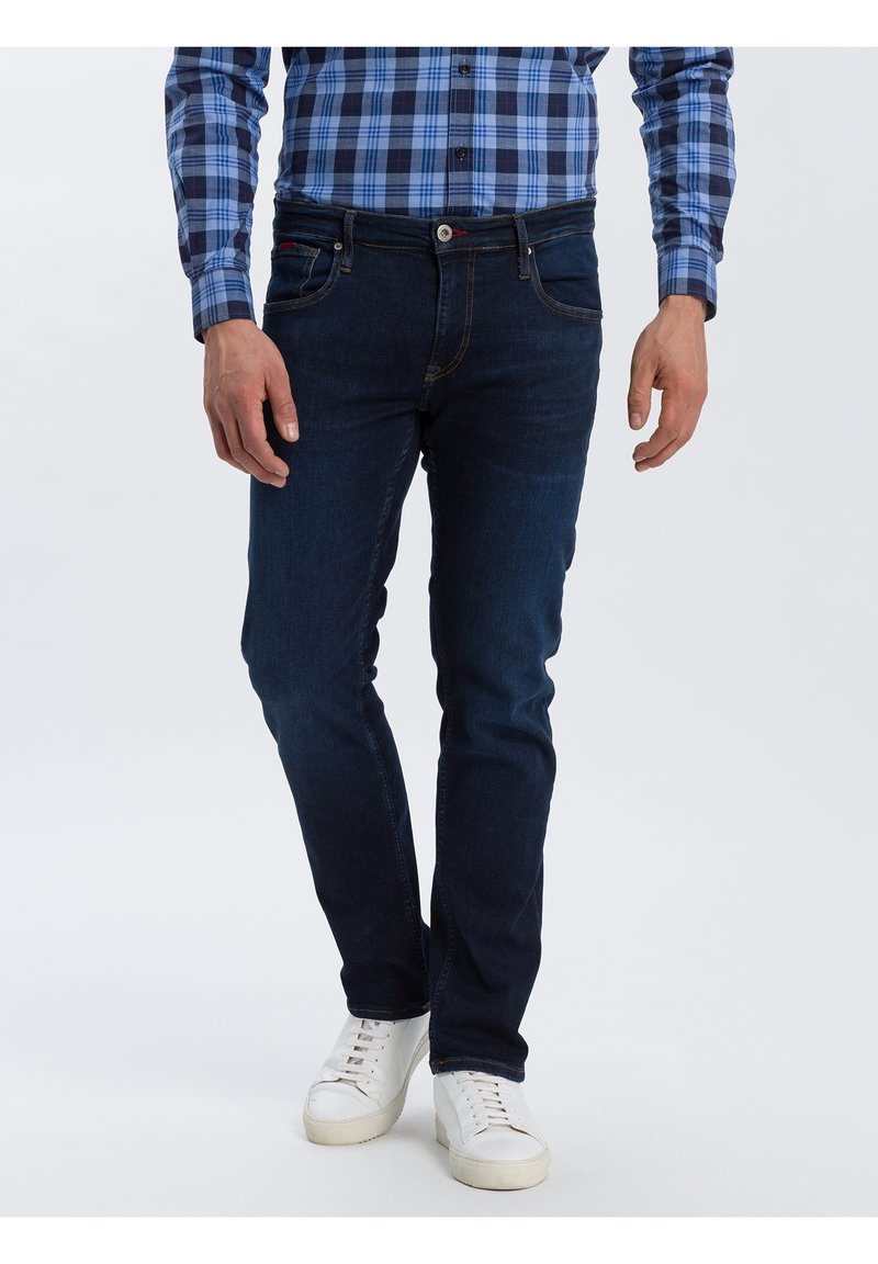 Muž oblečený v tmavě modrých slim-fit džínách, bílých teniskách a modré kostkované košili s dlouhým rukávem, stojící na jednolitém pozadí.