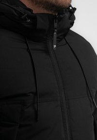 Giacca nera con cappuccio, dotata di chiusura con zip, corde regolabili e un tessuto liscio e strutturato con dettagli sulla tasca frontale.