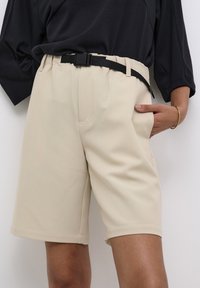 Pantalones cortos beiges hechos de una tela suave, con bolsillos laterales, una cinturilla con pasadores para cinturón y un cinturón negro. El diseño tiene un ajuste relajado.