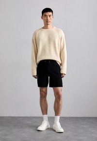 Cremefarvet strikket sweater med rund halsudskæring, parret med sorte shorts og hvide sneakers. Bemærkelsesværdige detaljer inkluderer ribstrikkede manchetter og en snoretræk i taljen.