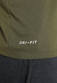 Närbild av en olivgrön Dri-FIT-topp över mörkgrå byxor, med fokus på den sydda fållen och den synliga DRI-FIT-logotypen.
