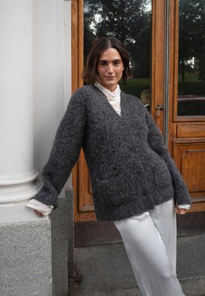 Dunkelgrau, flauschige Strickjacke mit V-Ausschnitt, Knopfverschluss und Fronttaschen; getragen über einem hellen Rollkragenpullover und kombiniert mit weißen Satin-Hosen.