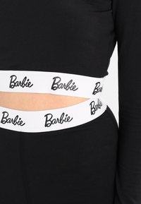 Svart åtsittande outfit med ett brett vitt elastiskt midjeband. "Barbie" är tryckt i svarta kursiva bokstäver längs midjebandet. Slät textur.