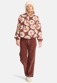 Brun fleece-tröja med ett stort mönster av vita och orangea prästkragar, halv dragkedja och elastiska ärmslut, kombinerat med rostfärgade kordbyxor och krämfärgade sneakers.