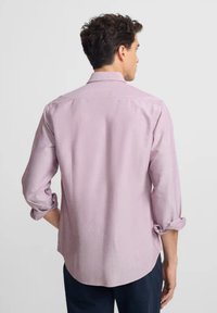 Camisa de manga larga color rosa claro con cuello abotonado, puños enrollados y una textura suave. Presenta un dobladillo curvado y detalles mínimos.