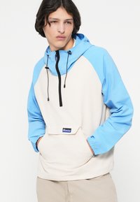 Leichte Pulloverjacke mit beigem Körper und blauen Ärmeln, ausgestattet mit einer Vordertasche, verstellbarem Kapuze und schwarzen Kordeln.