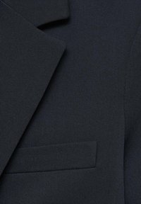 Chaqueta de traje azul marino de tejido fino, con solapa y un bolsillo en el pecho. Líneas limpias y diseño minimalista.