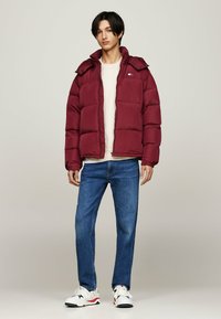 Jachetă puf burgundă cu glugă, având un design matlasat, mansete elastice, închidere cu fermoar, purtată peste o cămașă ușoară și blugi albaștri.