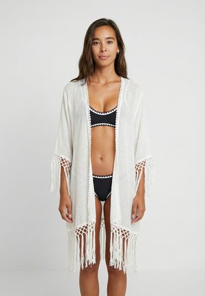 Biała narzutka z frędzlami na rękawach, wykonana z lekkiego materiału. Posiada misterny wzór szydełkowy na mankietach i dolnym brzegu. Nosi się ją na czarny bikini.