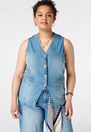 Vrouw draagt mouwloze denimvest en spijkerbroek met gestreepte sjaal hangend aan de taille tegen een witte achtergrond.