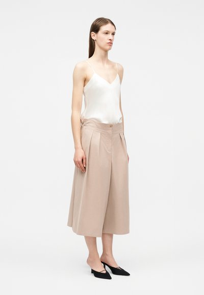 Γυναίκα που στέκεται, φοράει λευκή σατέν καμισόλα, μπεζ παντελόνι φαρδύ με cropped τελείωμα και μαύρα μυτερά παπούτσια με χαμηλό τακούνι σε λευκό φόντο.