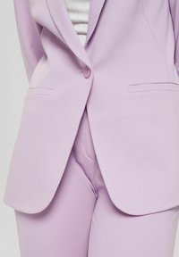 Veste blazer violet clair avec col cranté, fermeture à bouton et poches latérales, associée à un pantalon assorti sur mesure dans le même tissu et la même couleur.