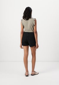 Olivenfarget ermeløs topp med volangskuldre, kombinert med høyt liv svarte denimshorts. Fottøy: beige sandaler. Bakgrunnen er nøytral.