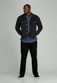 Moški nosi črn-denim jakno, modro srajco s ovratnikom, črne hlače in črne superge, stoji s rokami v žepih in gleda levo.