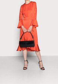Robe en satin orange avec des manches évasées et un ourlet volanté, associée à un sac à épaulette noir et des talons noirs à lanières.