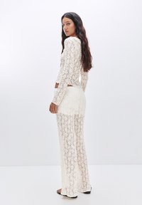 Weißes Spitzen-Zweiteiler-Set bestehend aus einem langärmeligen Oberteil und fließenden weiten Hosen, mit aufwendigen floralen Mustern und einer semi-transparenten Textur.