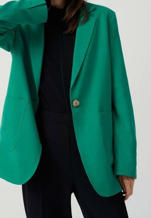 Personne portant un blazer vert à un seul bouton sur une tenue noire, debout devant un fond clair uni, avec un bras légèrement levé.