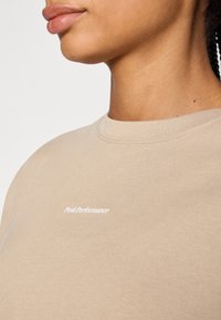 Beige katoenen t-shirt met een ronde hals, met een wit "PeakPerformance" logo op de borst, een gladde textuur en korte mouwen.