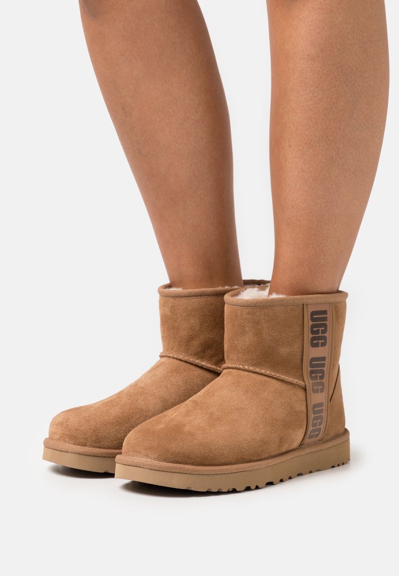 Personne portant des bottines UGG en daim beige avec intérieur duveteux et brides de marque sur fond blanc.