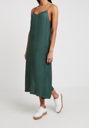 Žena v tmavě zelených midi šatech bez rukávů s tenkými ramínky a bílými platformovými teniskami, stojící před jednobarevným pozadím.