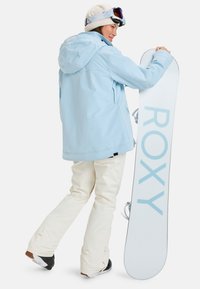 Hellblaues wasserdichtes Jacke mit Kapuze, cremefarbene Schneehose und schwarze Stiefel. In einer Hand wird ein weißes Snowboard mit blauem "ROXY"-Logo gehalten.