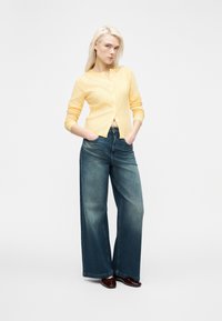 Ξανθιά γυναίκα στέκεται με τα χέρια στις τσέπες, φορώντας κίτρινο ζακέτα με κουμπιά, ξεβαμμένα φαρδιά μπλε τζιν και σκούρα λαμπερά παπούτσια σε λευκό φόντο.