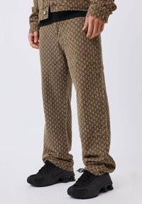 Pantaloni marroni a motivo con un tessuto testurizzato, caratterizzati da un design ripetuto. Abbinati a scarpe sportive nere.