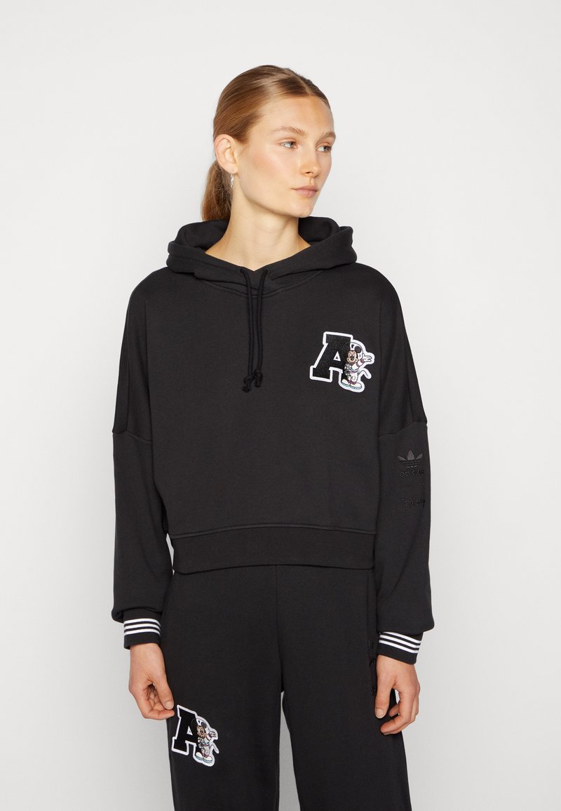 Black cropped hoodie made of soft material, featuring a front embroidered logo with a character, and striped cuffs for added detail.
