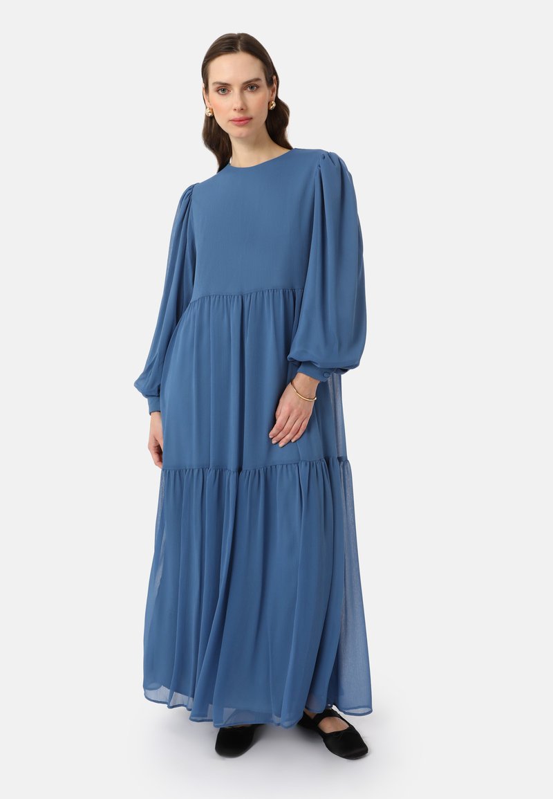Long, flowy blue dress made of lightweight fabric with a tiered design, round neckline, and puffed sleeves. Worn with black flats.