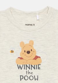 Svijetlo siva majica kratkih rukava s crtanom medvjedićem u crvenoj boji i natpisom "WINNIE the POOH", dopunjena malom ilustracijom pčele.