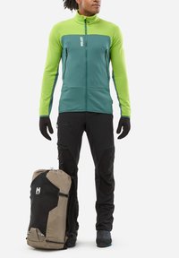 Person wearing a green and teal jacket with black gloves and pants standing next to a tan and black Millet backpack on a white background.