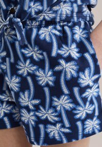 Mėlyni medvilniniai šortai su baltais palmės medžių raštais, turintys elastingą juosmenį ir užtrauktuko detales.