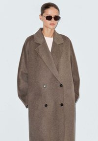Cappotto doppiopetto in misto lana marrone con revers a punta, vestibilità oversized e bottoni neri. Indossato con occhiali da sole rettangolari scuri.