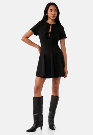 Vrouw in een korte zwarte jurk met strikdetails aan de voorkant en zwarte leren laarzen tot aan de knie, staand met één hand op de heup.